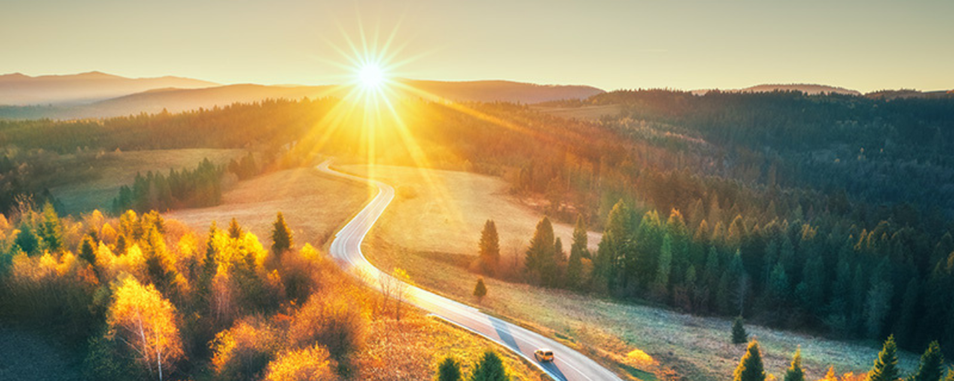 Car driving on road in wooded area at sunrise.