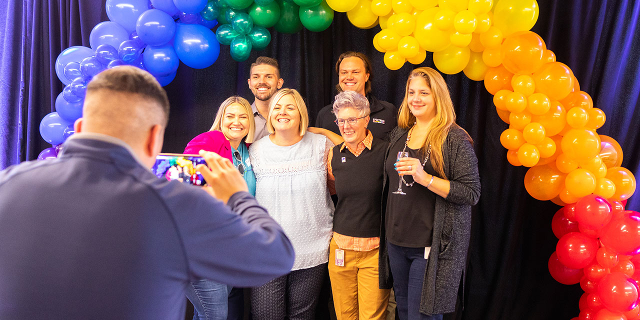 A group of Nexus associates pose for a photo under an arch of rainbow colored balloons at a corporate event