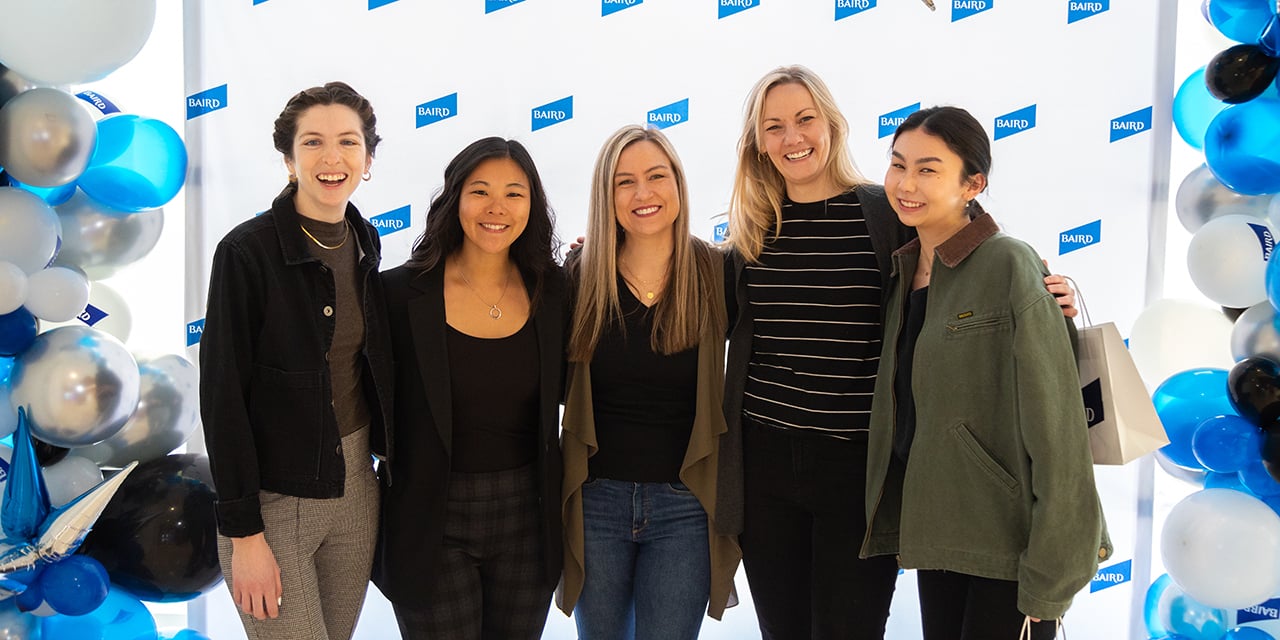 Five women stand before a Nexus backdrop with blue, white, and silver balloons.