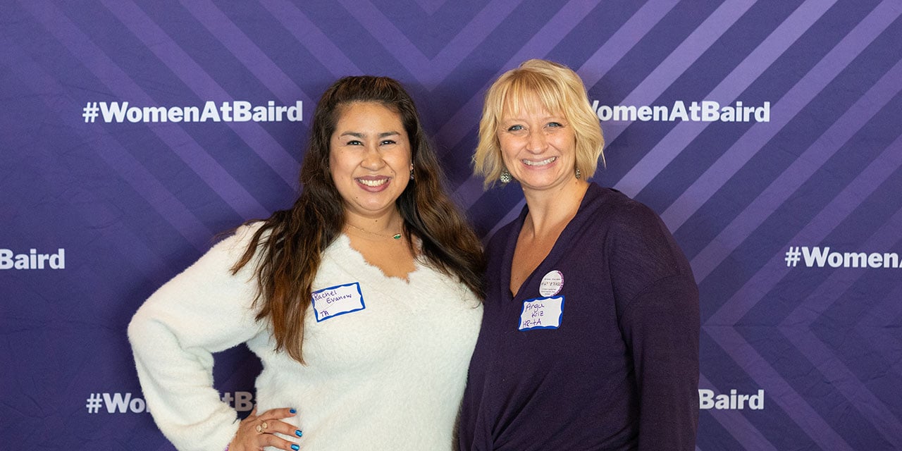 Two Nexus associates in front of a purple banner with #womenatbaird hashtags