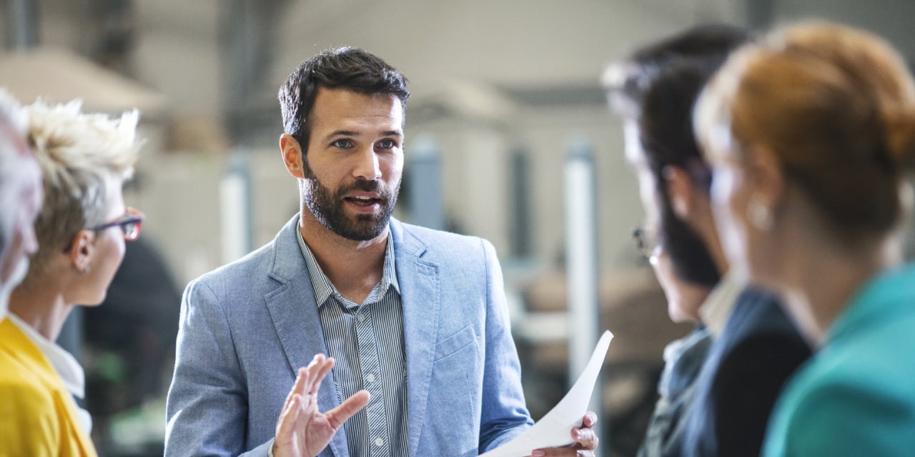 Five people in discussion indoors; central figure in blue blazer speaks while holding papers.